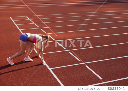 At start line. One Caucasian woman, female athlete, runner training at public stadium, sport court or running track outdoors. Summer sport games. 80835041