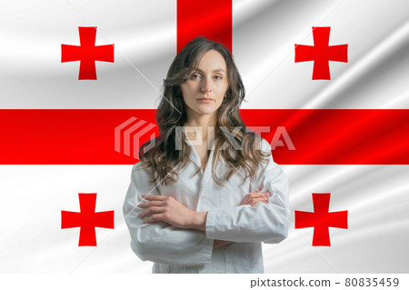 Medicine in Georgia. Happy beautiful female doctor in medical coat standing with crossed arms against the background of the flag of Georgia 80835459