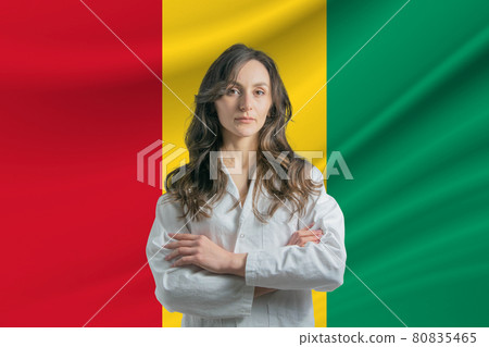 Medicine in Guinea. Happy beautiful female doctor in medical coat standing with crossed arms against the background of the flag of Guinea 80835465