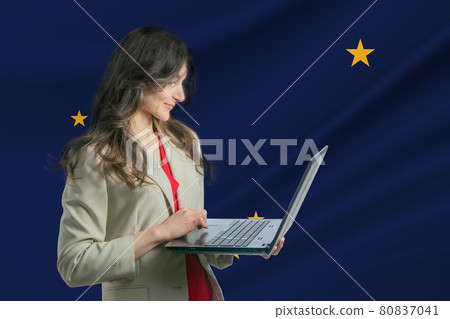 Freelance in Alaska. Beautiful young woman freelancer uses laptop computer against the background of the flag of Alaska Freelance in Alaska. Beautiful young woman freelancer uses laptop computer against the background of the flag of Alaska 80837041