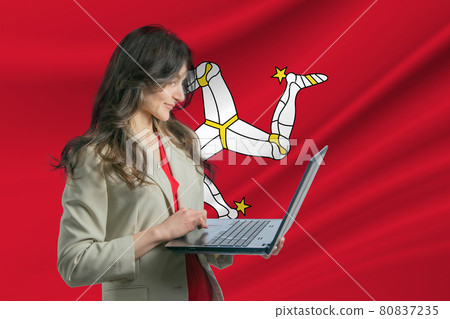 Freelance in Isle of Man. Beautiful young woman freelancer uses laptop computer against the background of the flag of Isle of Man Freelance in Isle of Man. Beautiful young woman freelancer uses laptop computer against the background of the flag of Isle of Man 80837235