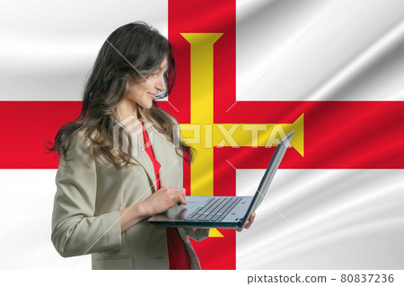 Freelance in Guernsey. Beautiful young woman freelancer uses laptop computer against the background of the flag of Guernsey Freelance in Guernsey. Beautiful young woman freelancer uses laptop computer against the background of the flag of Guernsey 80837236
