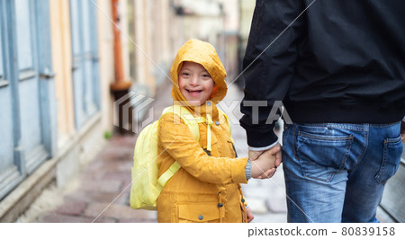 Happy down syndrome boy with unrecognizable father outdoors on a walk in rain, looking at camera. 80839158