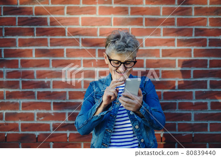 Senior woman standing outdoors against brick wall background, using smartphone. Senior woman standing outdoors against brick wall background, using smartphone. 80839440