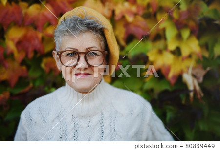 Senior woman standing outdoors against colorful natural autumn background, looking at camera. 80839449