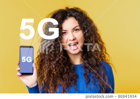 5G network. Wireless communication. Mobile Internet. ICT digital future. Innovative technology. Excited woman showing smartphone with white icon hologram isolated on orange. 5G network. Wireless communication. Mobile Internet. ICT digital future. Innovative technology. Excited woman showing smartphone with white icon hologram isolated on orange. 80841934