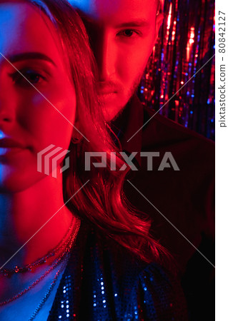 Party couple. Xmas portrait. Neon light people. Romantic festive night. Half face of confident glamour woman with bf in red blue glow on dark shiny tinsel. 80842127