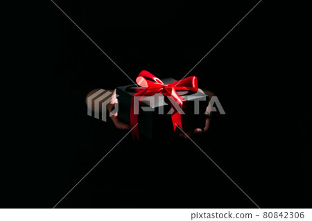 Christmas present. Festive surprise. Stylish decor. Unrecognizable woman holding elegant black gift box red ribbon in hands isolated dark light glow defocused. 80842306