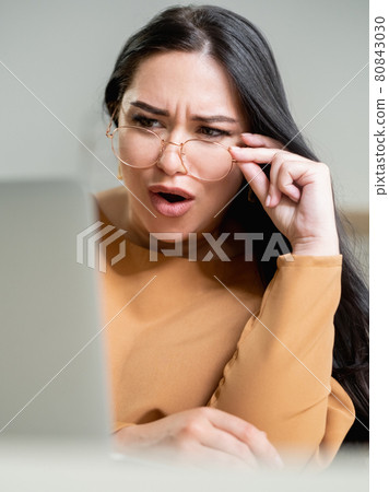 Shock content. Surprised woman. Unexpected situation. Unbelievable behavior. Pretty brunette lady touching spectacles looking on laptop in light room interior. 80843030