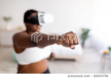 Virtual training. Overweight black lady in VR glasses using fighting simulator, making punch at home, focus on fist 80844796
