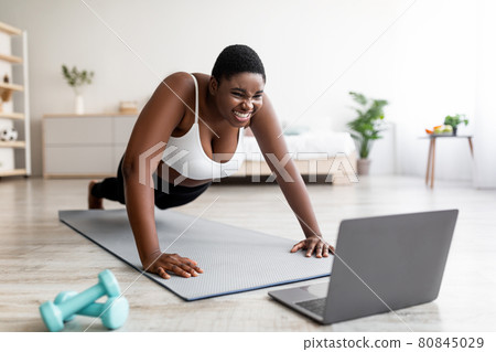 Plus size Afro woman standing in plank pose or doing push ups near laptop, working out to online sports tutorial at home Plus size Afro woman standing in plank pose or doing push ups near laptop, working out to online sports tutorial at home 80845029