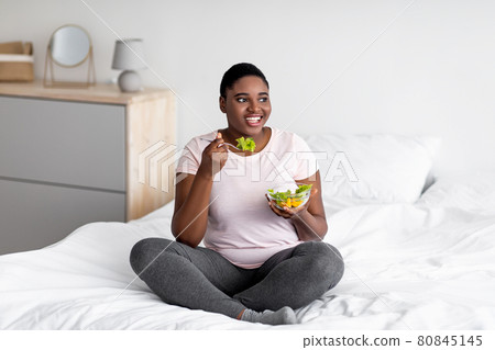 Balanced weight loss diet concept. Plus size Afro woman eating vegetable salad, sitting on bed at home 80845145