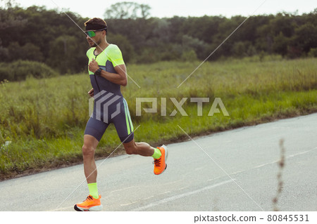 Young professional male triathlete, runner in motion on open road. Concept of sport, lifestyle, activity Young professional male triathlete, runner in motion on open road. Concept of sport, lifestyle, activity 80845531