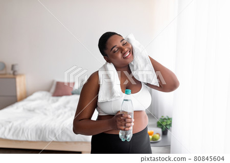 Plus size black woman with bottle of water wiping sweat with towel after domestic workout, indoors Plus size black woman with bottle of water wiping sweat with towel after domestic workout, indoors 80845604