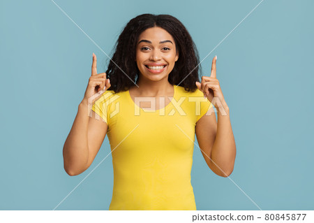 Look up. Excited african american lady pointing fingers upward showing something posing standing over blue background 80845877