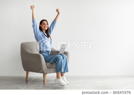 Ecstatic woman sitting in armchair with laptop, lifting hands up, making YES gesture, celebrating online win or success 80845976
