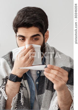 Seasonal infectious disease. Sick arab guy sneezing, measuring body temperature, sitting at home interior, crop, closeup Seasonal infectious disease. Sick arab guy sneezing, measuring body temperature, sitting at home interior, crop, closeup 80846004