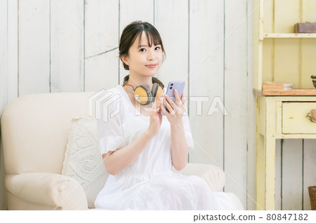 坐在沙發上,通過耳機聽音樂的年輕女人 坐在沙發上,通過耳機聽音樂的年輕女人 80847182