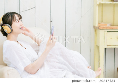 坐在沙發上,通過耳機聽音樂的年輕女人 坐在沙發上,通過耳機聽音樂的年輕女人 80847183