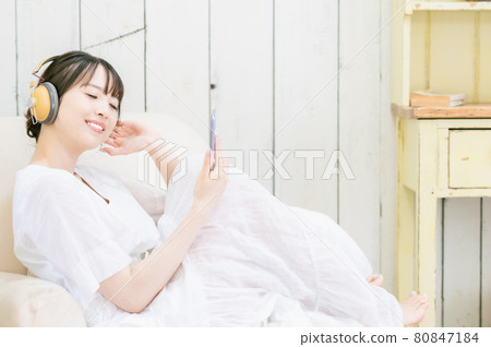 坐在沙發上,通過耳機聽音樂的年輕女人 坐在沙發上,通過耳機聽音樂的年輕女人 80847184