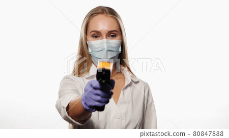 Doctor or nurse pointing sensor thermometer on camera wearing protective mask white medical hospital robe. Young blond haired nurse checking on patients or coworker temperature. 4k UHD Doctor or nurse pointing sensor thermometer on camera wearing protective mask white medical hospital robe. Young blond haired nurse checking on patients or coworker temperature. 4k UHD 80847888