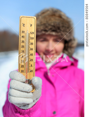 Young woman in pink jacket holds wooden thermometer outside on sunny but cold day, illustrating weather with freezing temperatures as low as -8 degrees Celsius. Young woman in pink jacket holds wooden thermometer outside on sunny but cold day, illustrating weather with freezing temperatures as low as -8 degrees Celsius. 80849564