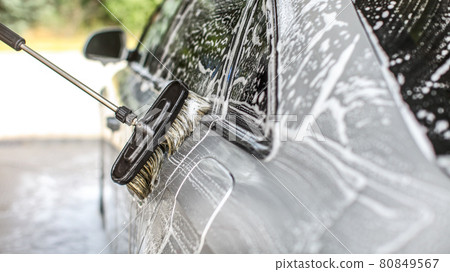 Side of silver car washed in self service carwash, detail on brush moving over white shampoo, blurred sun lit trees in background 80849567