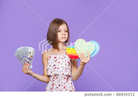 A little girl in a summer dress holds pop it in one hand, US dollar cash in the other and thinks. A little girl in a summer dress holds pop it in one hand, US dollar cash in the other and thinks. 80850151
