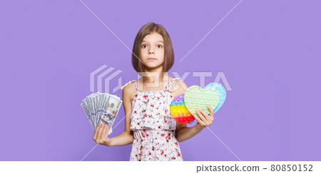 A little girl in a summer dress holds pop it in one hand, US dollar cash in the other and looking at camera A little girl in a summer dress holds pop it in one hand, US dollar cash in the other and looking at camera 80850152