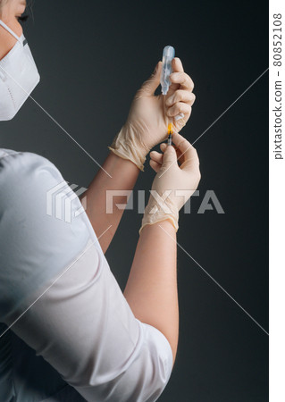 Close-up back view of nurse in white gloves and face mask holding and filling up vaccine to syringe on black isolated background. Close-up back view of nurse in white gloves and face mask holding and filling up vaccine to syringe on black isolated background. 80852108