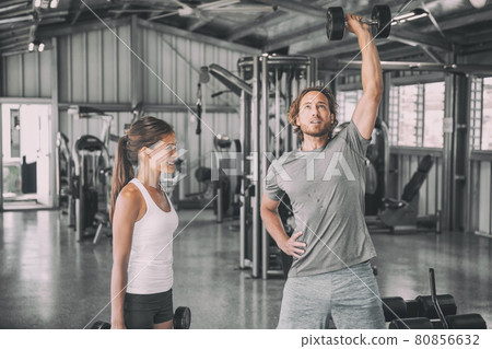 Fitness gym personal trainer coach training Asian woman with free weights dumbbell bicep curls workout talking having fun exercise partner. Fit man, happy girl. Fitness gym personal trainer coach training Asian woman with free weights dumbbell bicep curls workout talking having fun exercise partner. Fit man, happy girl. 80856632