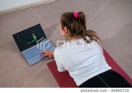 A chubby young woman is watching an online fitness lesson on a laptop. Distance sports training. A chubby young woman is watching an online fitness lesson on a laptop. Distance sports training. 80860728
