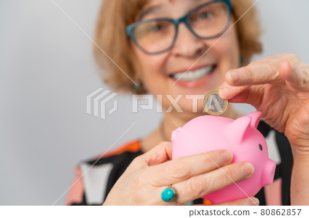 An elderly woman with glasses puts a coin in a piggy bank on a white background 80862857