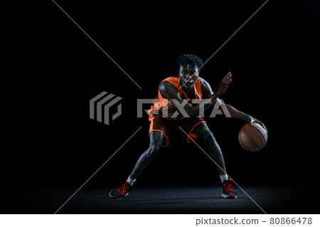 Front view. Young african man, basketball player practicing with ball isolated over dark studio background. 80866478