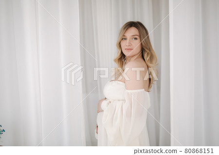Beautiful pregnant woman in elegant white dress posing to photographer in studio. Background of white tulle Beautiful pregnant woman in elegant white dress posing to photographer in studio. Background of white tulle 80868151