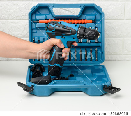 a female hand holds a portable drill on a battery against the background of an open plastic suitcase with attachments 80868616