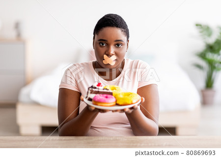Hungry plus size black woman having weight loss diet, wearing adhesive bandages on mouth, holding plate with sweets 80869693
