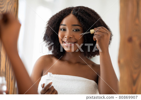 Smiling young black lady in towel using facial serum near mirror, taking care about her beauty at home 80869907