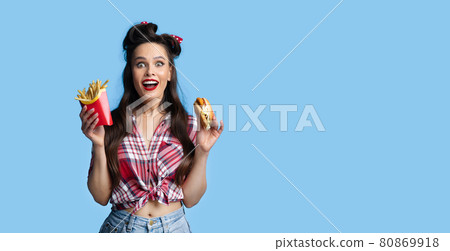 Portrait of excited young pinup lady in retro outfit holding hamburger and french fries on blue background, banner Portrait of excited young pinup lady in retro outfit holding hamburger and french fries on blue background, banner 80869918