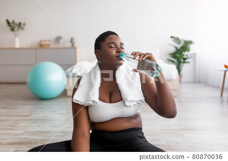 Wellness and slimming. Overweight black lady drinking clear sparkling water from bottle after domestic workout Wellness and slimming. Overweight black lady drinking clear sparkling water from bottle after domestic workout 80870036