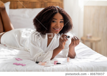 Gorgeous Afro lady doing manicure, applying nail polish, smiling at camera on bed at home. Domestic spa salon Gorgeous Afro lady doing manicure, applying nail polish, smiling at camera on bed at home. Domestic spa salon 80870037
