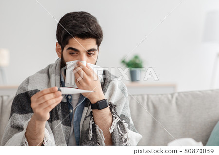Cold, flu, coronavirus concept. Sick arab man sitting on couch and sneezing, measuring body temperature, home interior Cold, flu, coronavirus concept. Sick arab man sitting on couch and sneezing, measuring body temperature, home interior 80870039