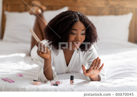 Pretty black woman lying on bed with nail file, doing manicure, taking care of her hands at home Pretty black woman lying on bed with nail file, doing manicure, taking care of her hands at home 80870045