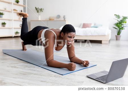 Plus size Afro lady working out at home with online personal trainer, using laptop, standing in elbow plank pose Plus size Afro lady working out at home with online personal trainer, using laptop, standing in elbow plank pose 80870138