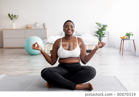 Overweight black woman sitting on sports mat in lotus pose, meditating, doing yoga practice at home 80870319