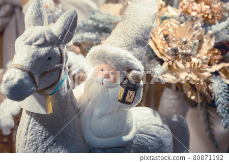Toy Santa Claus in a white fur coat riding a white horse with a lantern in his hand. 80871192