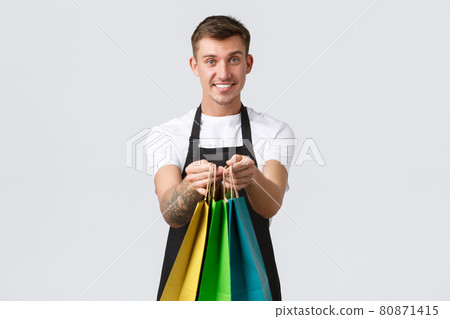 Retail store, shopping and employees concept. Friendly handsome and polite salesman handing over eco-bags with purchased items to client in shop, standing white background 80871415
