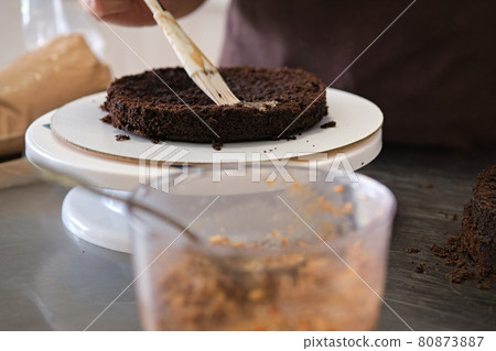 Woman pastry chef impregnates chocolate sponge cake on stand, close-up. Cake making process, Selective focus Woman pastry chef impregnates chocolate sponge cake on stand, close-up. Cake making process, Selective focus 80873887