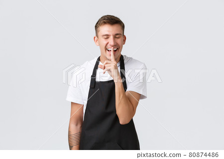 Small business owners, coffee shop and staff concept. Handsome happy barista, waiter in cafe wearing black apron, laughing and make shush gesture, asking keep quiet or not tell secret 80874486