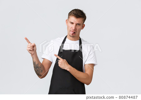 Small business owners, coffee shop and staff concept. Disappointed grimacing barista in black apron, pointing upper left corner, showing tongue from disgust or dislike, standing white background 80874487